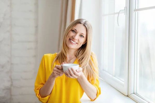 Vrolijke mooie jonge blonde Europese vrouw in gele kleren geniet van hete thee en vrije tijd in de buurt van venster — Stockfoto