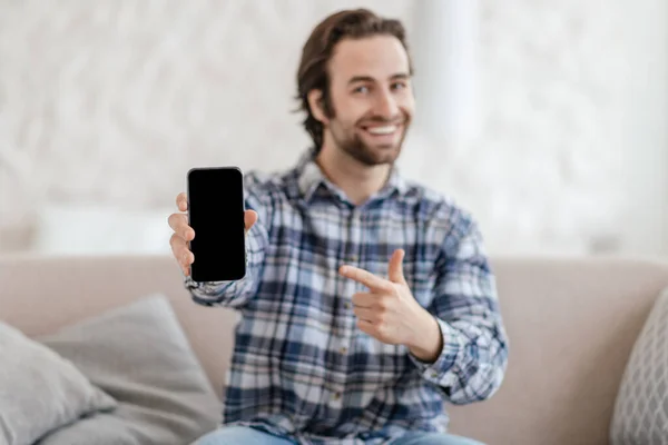 Zufriedene Millennial-Kaukasier mit Stoppeln sitzen auf dem Sofa und zeigen mit dem Finger auf Telefon mit leerem Bildschirm im Wohnzimmer — Stockfoto