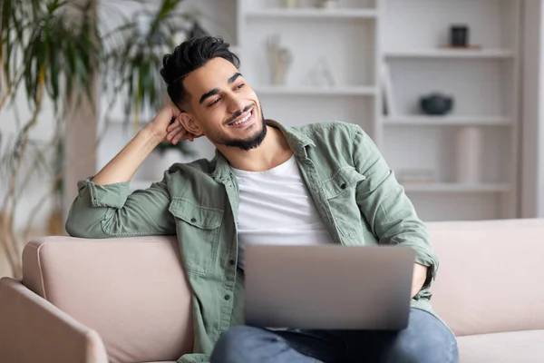 Lächelnder junger muslimischer Mann mit Bart wacht im freien Raum, sitzt auf Sofa mit Laptop im Wohnzimmer — Stockfoto