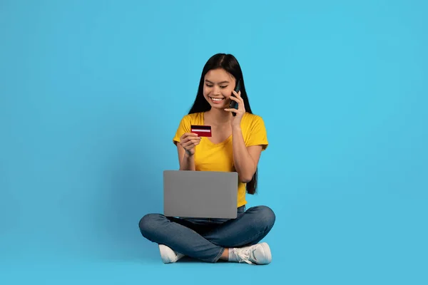 Cheerful young asian female in casual sitting on floor with laptop, calling phone and showing credit card —  Fotos de Stock