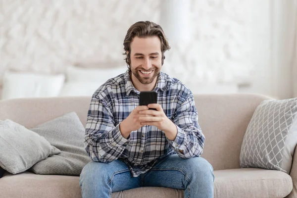 Smiling millennial caucasian guy with stubble sits on sofa typing on phone alone in living room interior — Stok fotoğraf