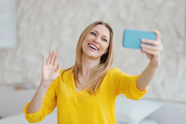 Lächelnde junge blonde Europäerin in gelben Kleidern winkt mit der Hand und blickt in die Smartphone-Kamera — Stockfoto