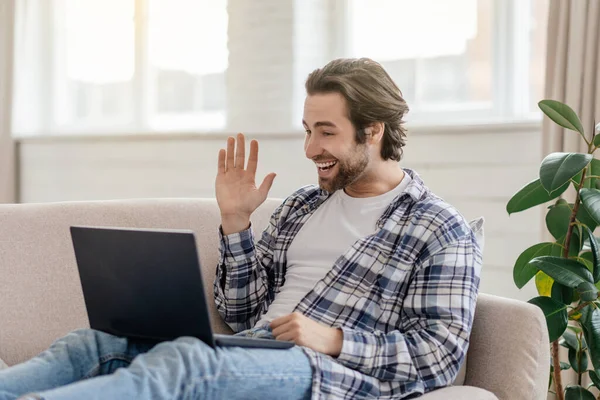 Happy millennial caucasian man with stubble sits on sofa waving hand and looking at laptop in living room — Foto de Stock