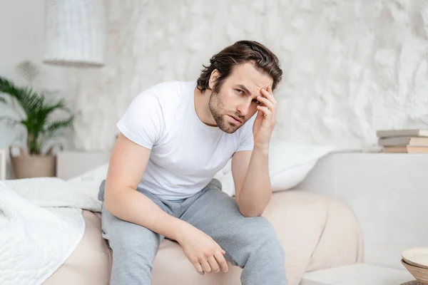 Sad millennial caucasian man with stubble sits on white bed, presses his hands to head and suffers from migraine — Foto de Stock