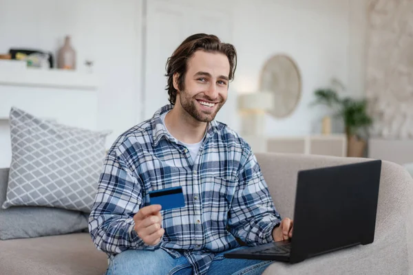 Vidám, millenniumi fehér fickó borostával ül a kanapén laptoppal és hitelkártyával a nappaliban — Stock Fotó