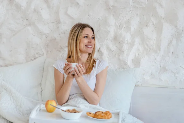 Frohe junge blonde Europäerin hält eine Tasse Kaffee in der Hand, frühstückt im Bett und blickt ins Leere — Stockfoto
