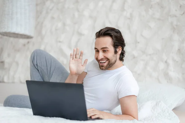Smiling handsome millennial caucasian guy with stubble lying on bed, waving hand at computer in bedroom — Foto de Stock