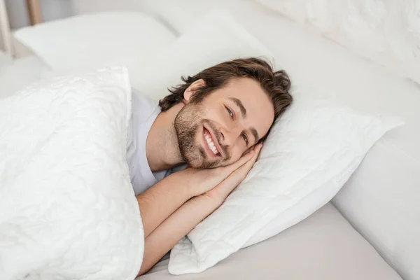 Smiling millennial caucasian male with stubble wake up, lies on white bed, enjoys free time and weekend — стоковое фото