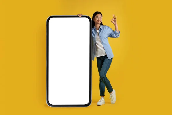 Satisfied young chinese woman in casual show ok sign with hand, stands near huge phone with empty screen — 图库照片