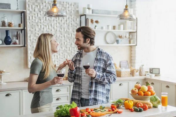Tatmin olmuş genç Avrupalı aile sohbetleri, modern mutfaktaki sebzelerle masada kadeh tokuşturmalar. — Stok fotoğraf
