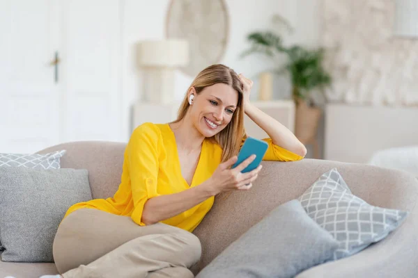 Cheerful young blonde european lady in yellow clothes with wireless headphones looks at phone, sits on sofa — Stockfoto