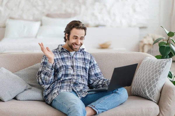 Satisfied millennial caucasian man with stubble sits on sofa waving hand and looking at laptop — Foto de Stock