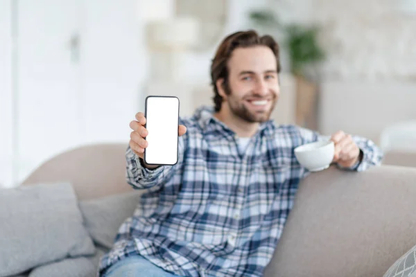 Sonriente joven caucásico hombre con rastrojo se sienta en el sofá con taza de café, muestra el teléfono con pantalla en blanco — Foto de Stock
