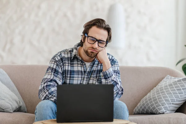 Tired young european male with stubble in glasses sits on sofa and looks at laptop in living room interior — Photo