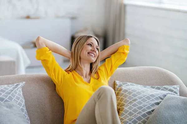 Satisfied young caucasian blonde woman in yellow clothes sits on sofa with hands behind head, rests on coach — Stok fotoğraf