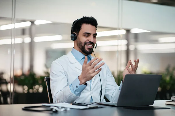 Smiling attractive millennial muslim guy doctor with beard in headphones, white coat looks at laptop — Foto de Stock