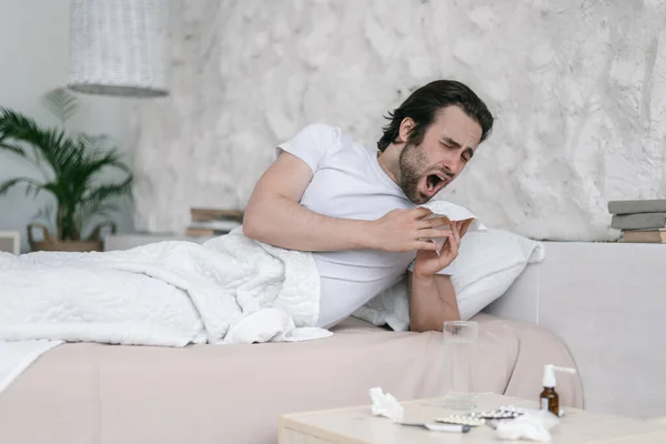 Θλιβερός λευκός άντρας χιλιετίας με γένια ξαπλώνει στο κρεβάτι, φτερνίζεται σε χαρτοπετσέτα στο εσωτερικό της κρεβατοκάμαρας με φάρμακα. — Φωτογραφία Αρχείου