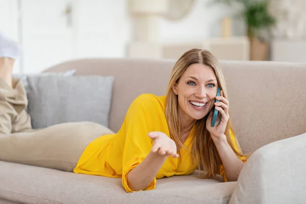 Smiling pretty millennial blond caucasian woman in yellow clothes speaks by phone, lies on sofa — Φωτογραφία Αρχείου