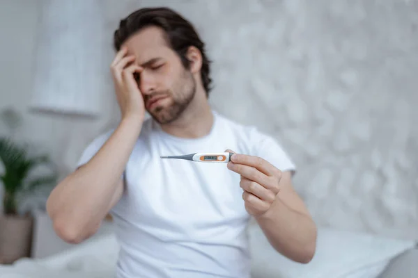Sad despaired millennial caucasian male with stubble, sits on bed presses his hand to head and holds thermometer — Stockfoto