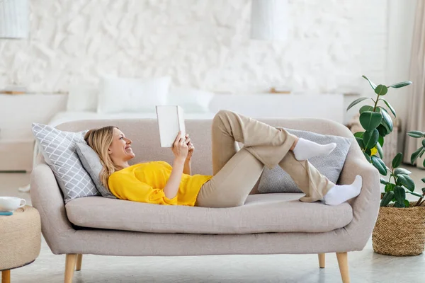 Ευτυχισμένος όμορφη νεαρή ξανθιά ευρωπαϊκή γυναίκα σε κίτρινα ρούχα βρίσκεται στον καναπέ και διαβάζει το βιβλίο — Φωτογραφία Αρχείου