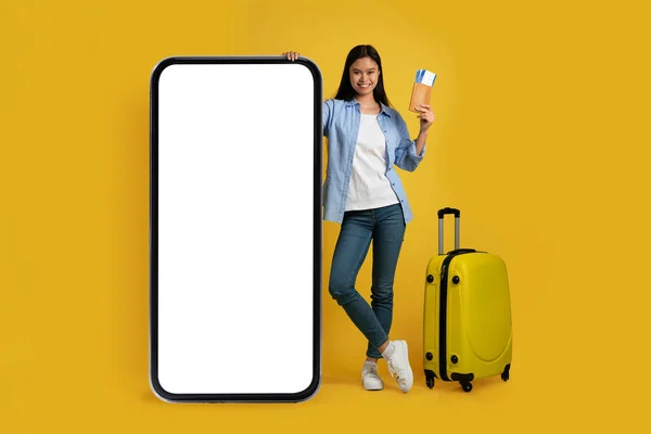 Cheerful young asian lady in casual with suitcase shows passport, air ticket stands near huge phone — 图库照片