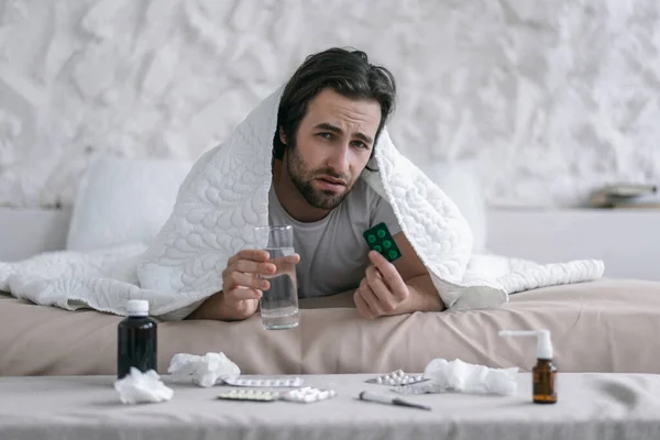 Unhappy millennial caucasian guy with stubble lies on bed under blanket holds glass of water and medicine — 图库照片