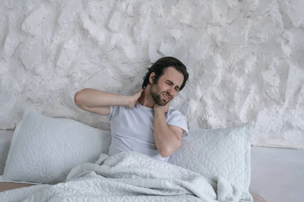 Δυσάρεστη χιλιετή θλίψη καυκάσιος τύπος με γένια ξυπνήσει, κάθεται στο κρεβάτι και υποφέρει από πόνο στους μυς του λαιμού — Φωτογραφία Αρχείου