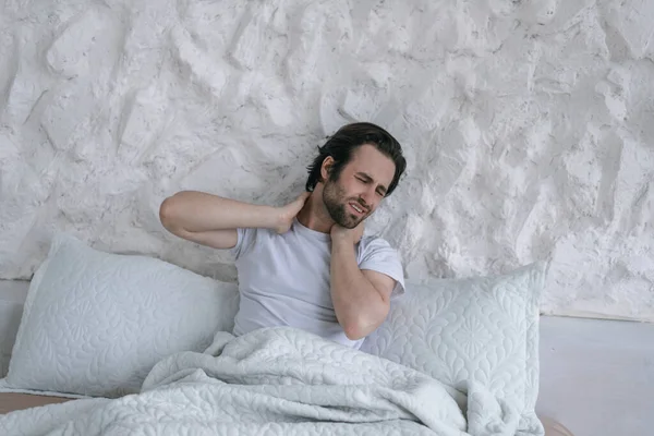 Unhappy sad millennial caucasian guy with stubble wake up, sitting on bed and suffering from pain in neck muscles