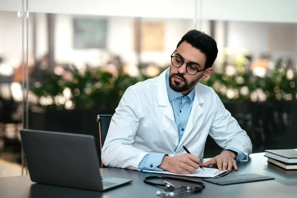 Serious attractive millennial arab guy doctor with beard in glasses, white coat looks at laptop, makes notes — Stock Fotó