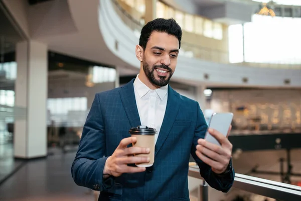Smiling handsome millennial islamic guy with beard in suit with cup of coffee takeaway looks at phone — Zdjęcie stockowe
