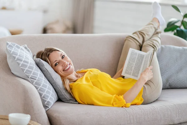 Smiling attractive millennial blond european lady in yellow clothes lies on sofa and reads book in living room — Φωτογραφία Αρχείου