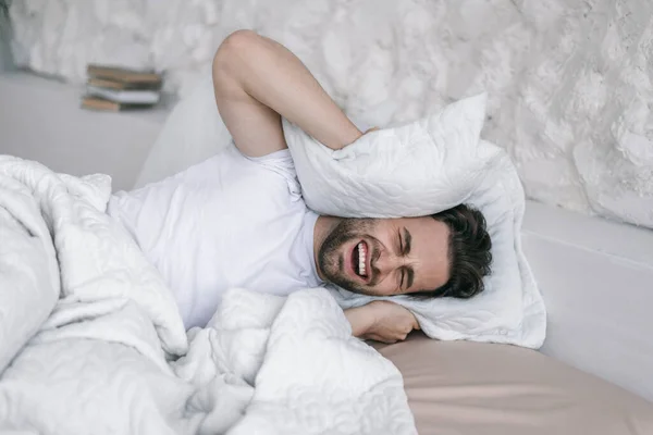 Despair millennial caucasian man with stubble covers ears with pillow on bed, suffers from noise and insomnia — Fotografia de Stock