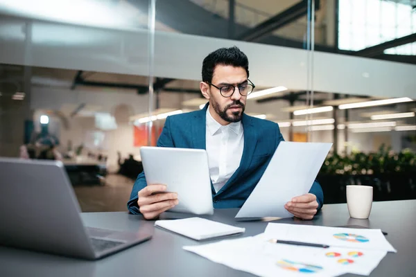 Serious busy handsome young muslim guy with beard in glasses, suit sits at table with computer and tablet — Stockfoto