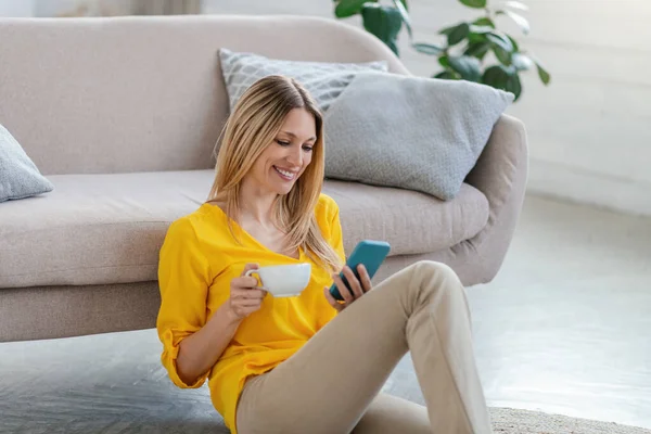 Smiling pretty young blond european lady in yellow clothes sits on floor, reads message on phone — Φωτογραφία Αρχείου