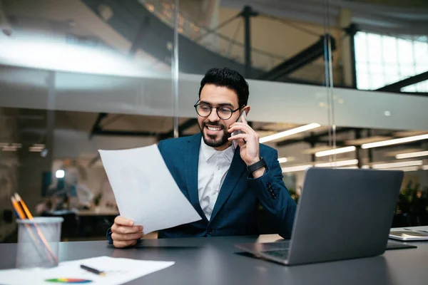 Alegre chico árabe joven guapo con barba en gafas, traje en la mesa con PC, llamada por teléfono inteligente — Foto de Stock