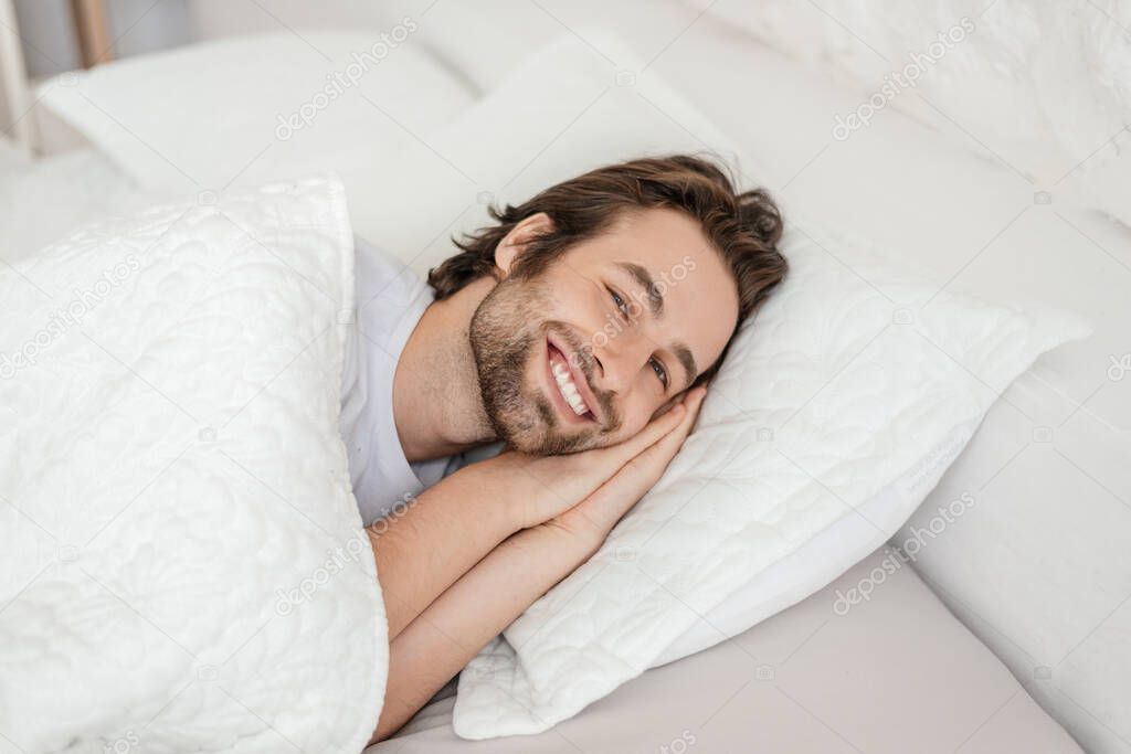 Smiling millennial caucasian male with stubble wake up, lies on white bed, enjoys free time and weekend