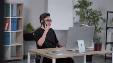 Gözlüklü genç adam bilgisayarla çalışıyor cep telefonuyla arkadaşlarıyla konuşuyor, iş yerinde oturuyor.