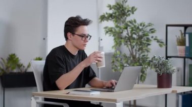 Gözlüklü genç bir öğrenci dizüstü bilgisayarda yazıyor ve dışarıdan kahve içiyor, evde çalışıyor, yavaş çekim, boş alan