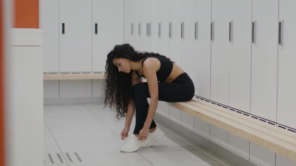 Sporty lifestyle. Young woman wearing sport suit tying laces on her sneakers, preparing for workout at gym locker room — Stockvideo