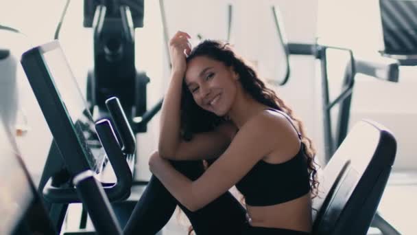 Beauty portrait of young gorgeous sporty lady sitting on training apparatus at gym and smiling to camera, slow motion — Stockvideo