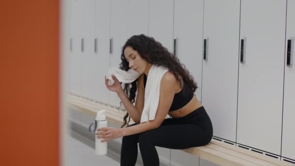 Young active sporty woman resting after hard workout at gym locker room, drinking water and wiping sweat from face — Stockvideo
