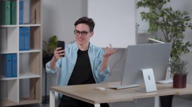 Genç pozitif adam arkadaşlarıyla akıllı telefondan sohbet ediyor, modern ofiste tek başına oturuyor, yavaş çekim, serbest alan