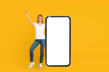 Beyaz tişörtlü gülümseyen genç Avrupalı kadın başparmağını kaldırıyor ve ekranı boş, kocaman bir telefonu var.