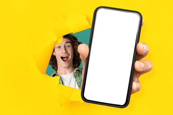 Glad excited young caucasian guy with open mouth looks through hole in yellow paper and shows smartphone — 스톡 사진
