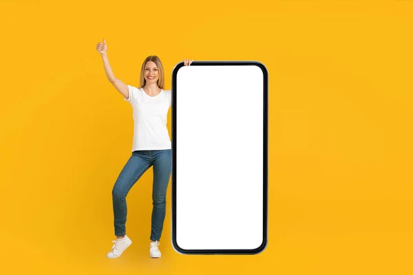 Smiling young european lady in white t-shirt showing thumb up stands with huge phone with empty screen — 图库照片