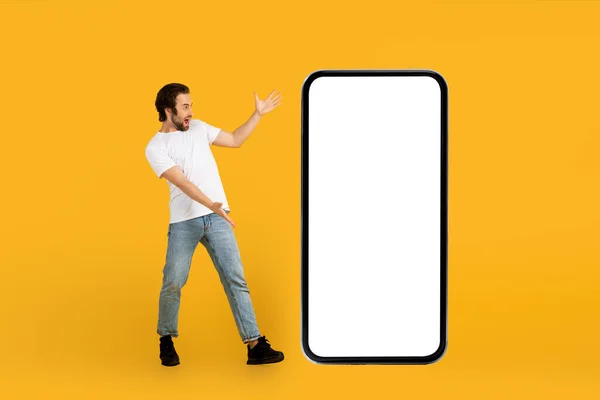 Cheerful shocked young european guy in white t-shirt looks in surprise at huge phone with blank screen — Stock fotografie