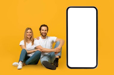 Neşeli genç Avrupalı çift yerde oturuyor. Kocaman akıllı telefonları ve boş ekranları var.