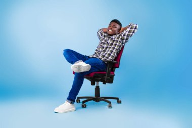 Yakışıklı, mutlu, genç, siyah bir adam. Mavi stüdyo arka planında, koltukta dinleniyor.