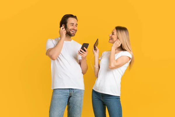 Happy millennial european male and female in white t-shirts with smartphones, puts headphones and listen music — Stockfoto