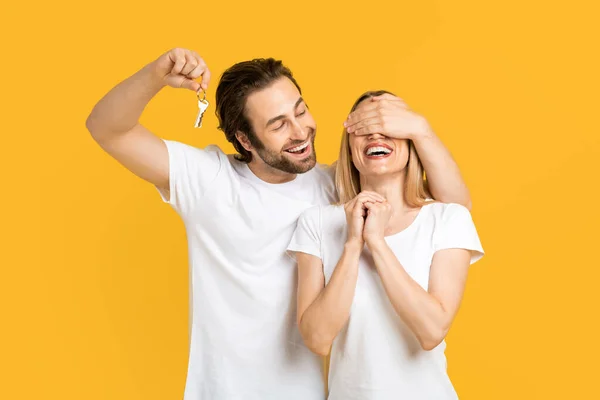 Smiling young european guy in white t-shirt closes eyes to his wife and holds keys, isolated on yellow background — Stok fotoğraf